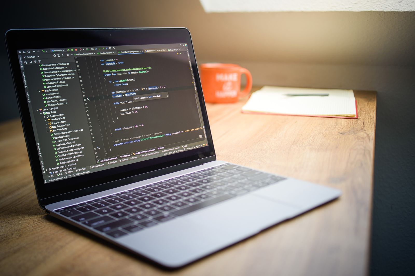Laptop displaying software code beside a notebook and a mug